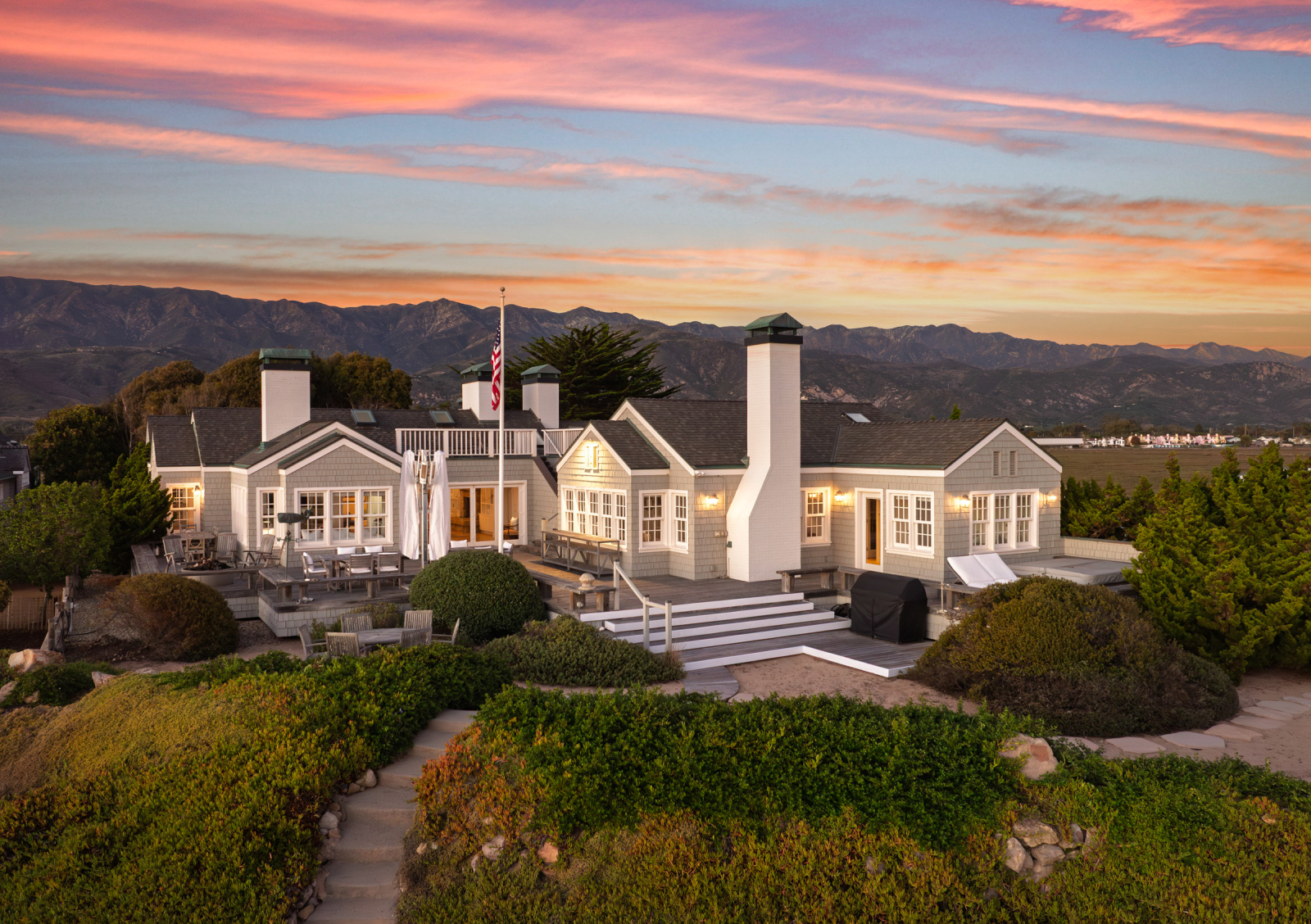 Coastal-style home with shingle siding and multiple chimneys, set on a landscaped bluff with expansive decking, outdoor seating, and sweeping mountain views under a colorful sunset sky.